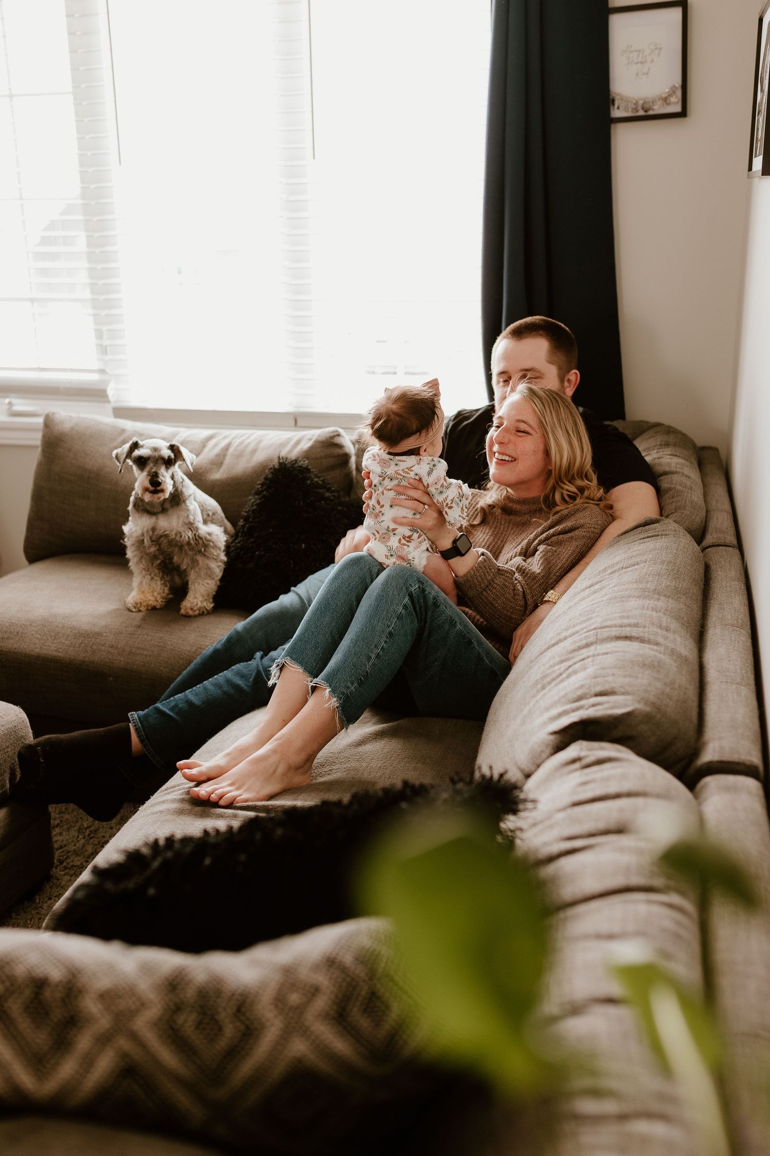 a new mom and dad sitting on a couch with their dog and new baby in their lap, smiling and playing with her