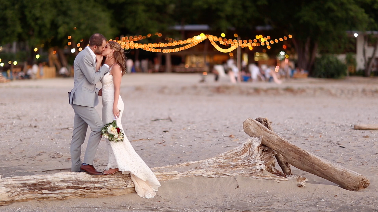 Romantic beach wedding photo with couple silhouetted against driftwood at sunset.