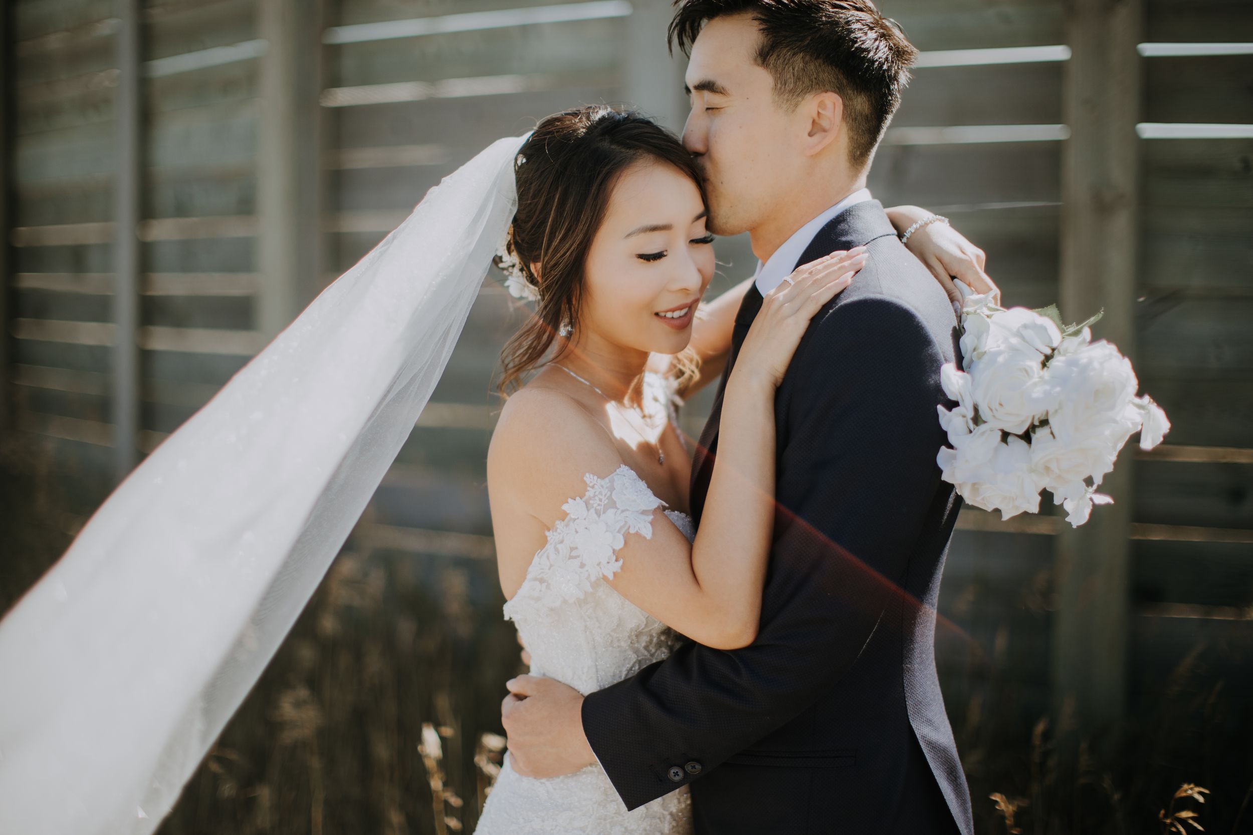 Elegant couple embraces during outdoor wedding photoshoot with flowing white veil and bouquet.