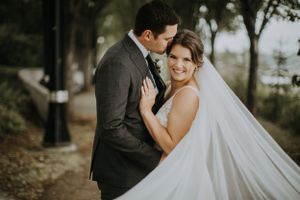 A romantic wedding couple embraces on a tree-lined path with a long flowing white veil trailing behind.