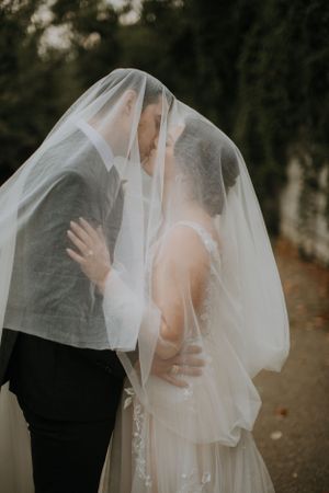 A romantic wedding photo shows a couple sharing a tender moment under a delicate white veil in an outdoor setting.