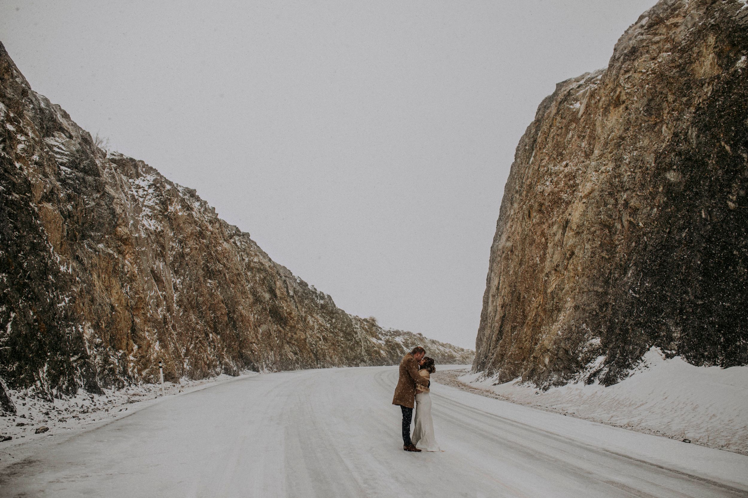 A person embraces their companion on a snowy mountain road between towering rocky cliffs.