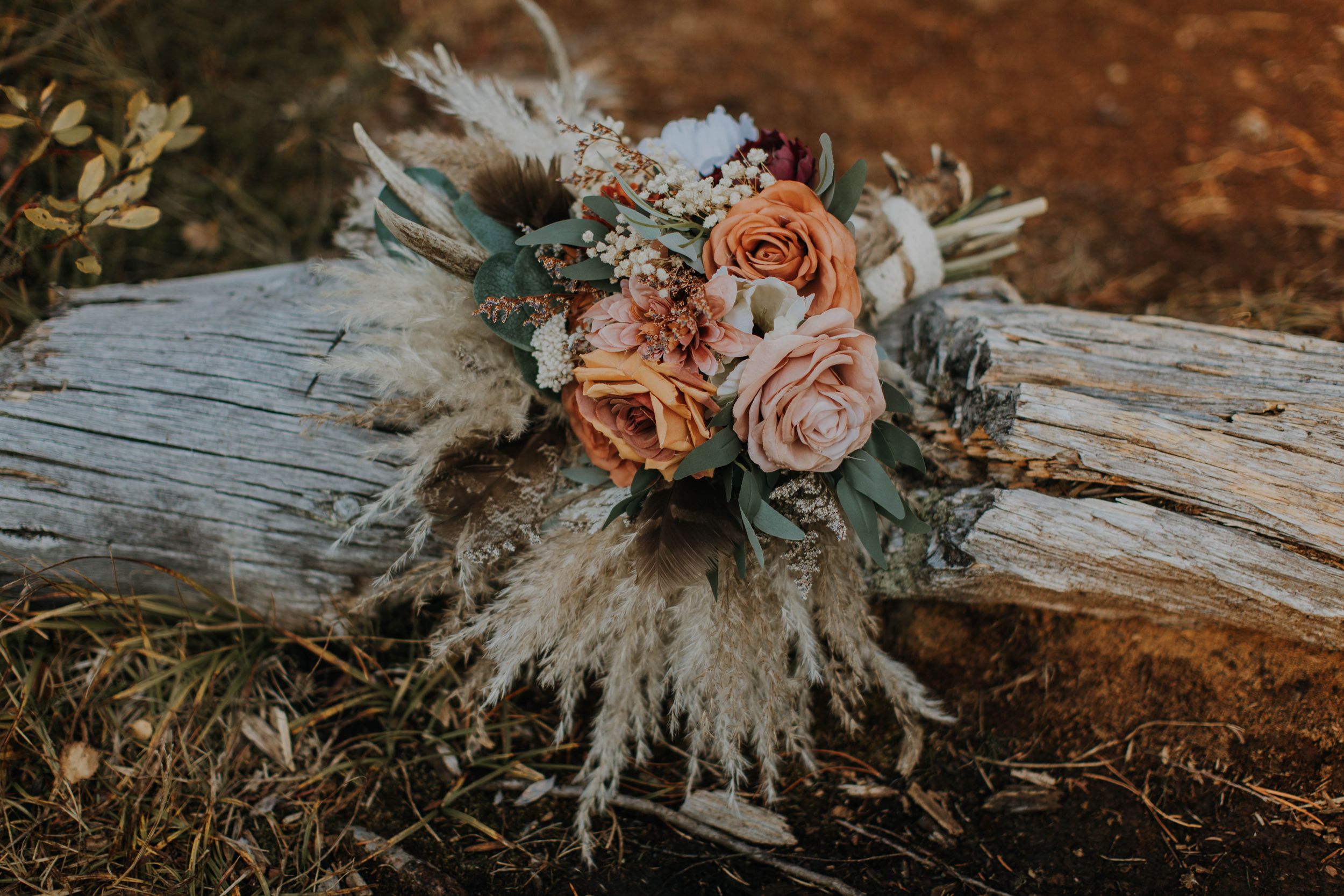 Rustic boho wedding bouquet with peach roses and pampas grass rests on weathered log in warm sunset light.