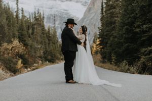 Newlyweds share intimate moment on mountain road with dramatic rocky peaks in background.