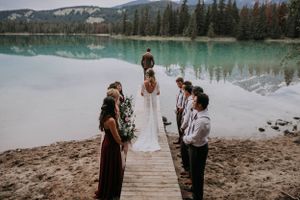 A small wedding ceremony takes place on a wooden dock overlooking a pristine turquoise mountain lake surrounded by evergreen forest.