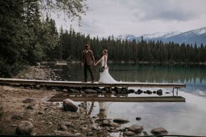 Newlywed couple standing on wooden dock at mountain lake surrounded by evergreen forest and snow-capped peaks.