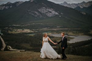 A couple in formal attire walks along a mountain ridge overlooking a scenic valley and mountain range.