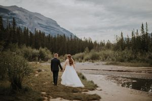 A couple holding hands walks along a river path with majestic mountain views in the Canadian Rockies wilderness.
