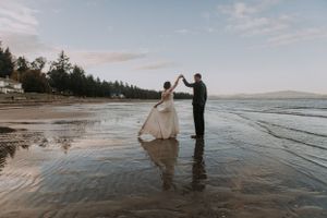 A romantic couple dancing on a serene beach at sunset with ocean waves and forest in the background.