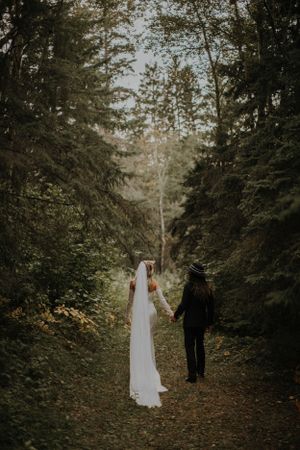 A couple in wedding attire walking hand in hand down a forest path surrounded by tall evergreen trees.