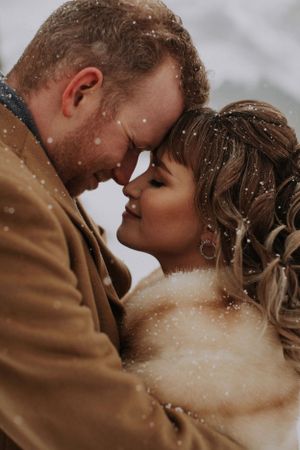 Intimate winter wedding moment captured with falling snow as couple embraces in warm coats.