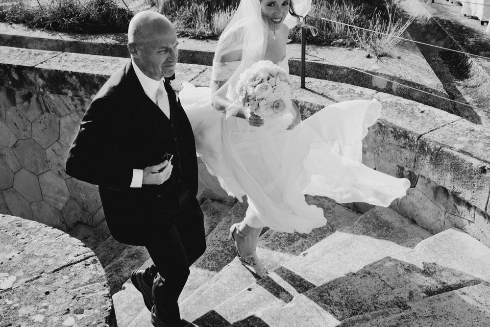Black and white photo of wedding party ascending stone steps with bride's veil flowing in the wind.