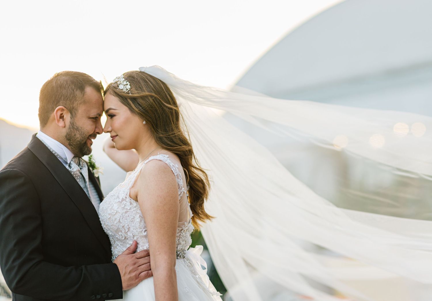 A couple embraces on their wedding day while a long white veil blows in the breeze against a soft, misty backdrop.