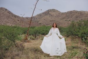 White gown billows in the breeze against rugged desert mountains and sparse vegetation.