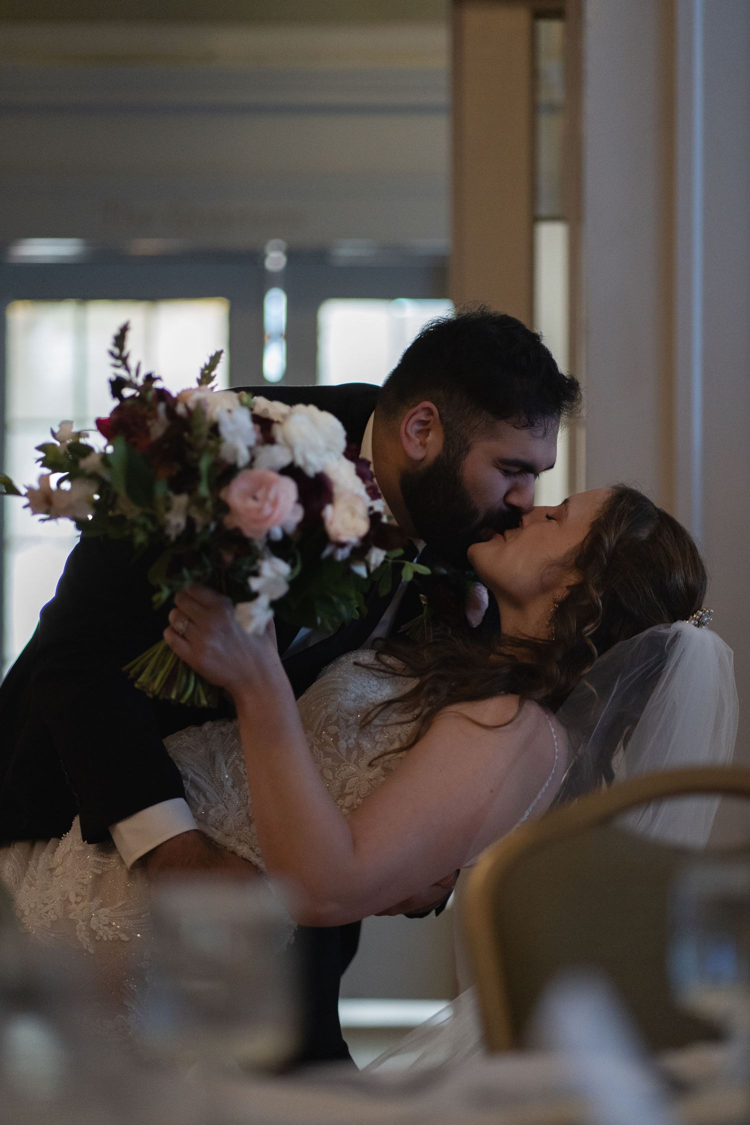 Romantic wedding couple sharing kiss while holding floral bouquet indoors.