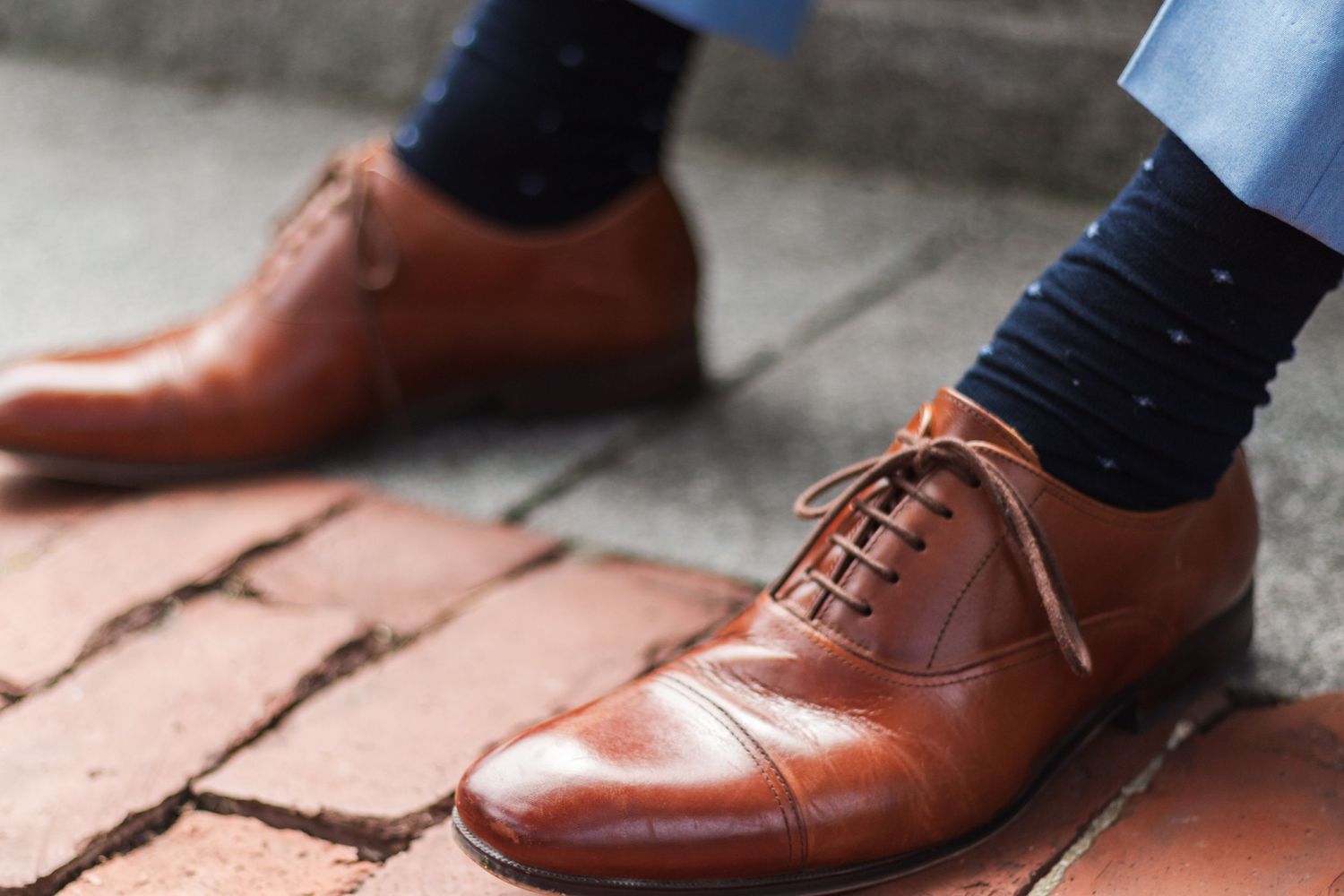 Brown leather oxford dress shoes with navy socks on brick pavement.