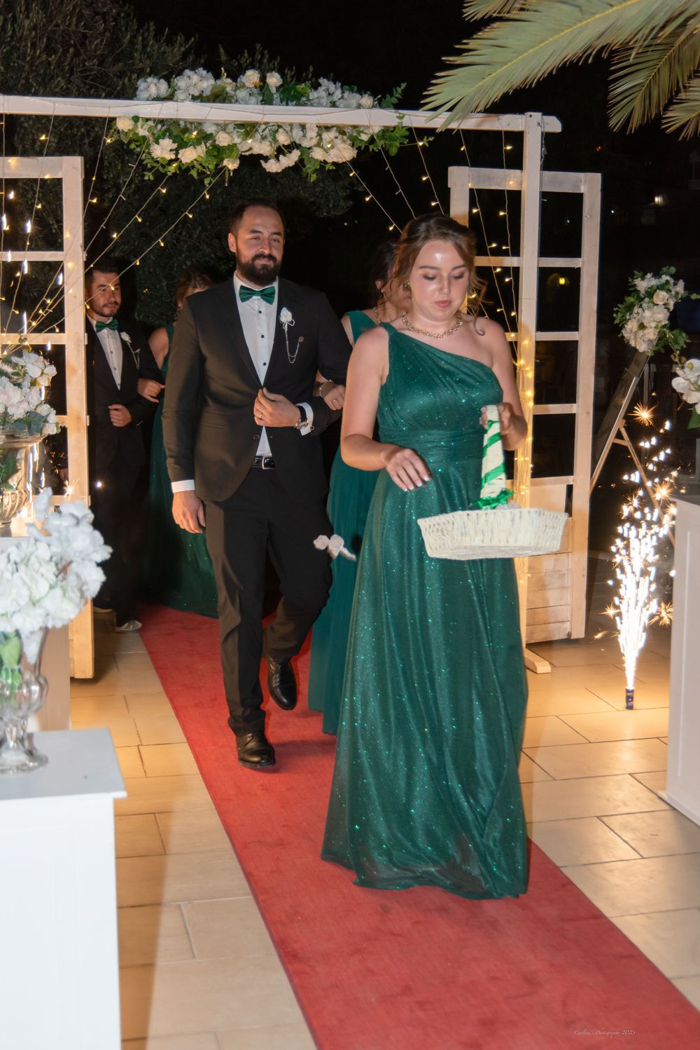 Wedding party members walking down red carpet under floral arch with sparkling lights.