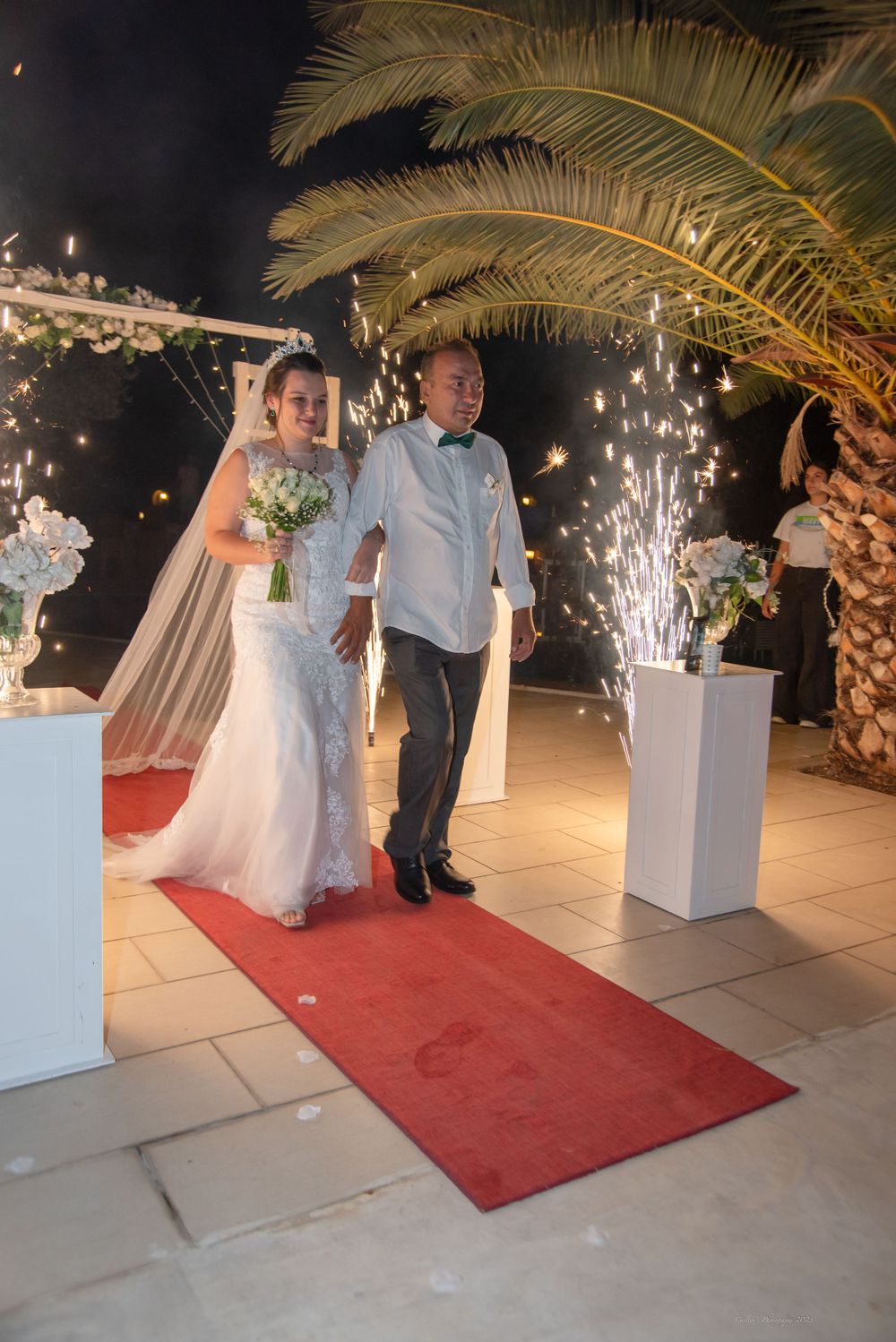 Couple walking down red carpet under floral arch with sparkler effects at nighttime wedding.