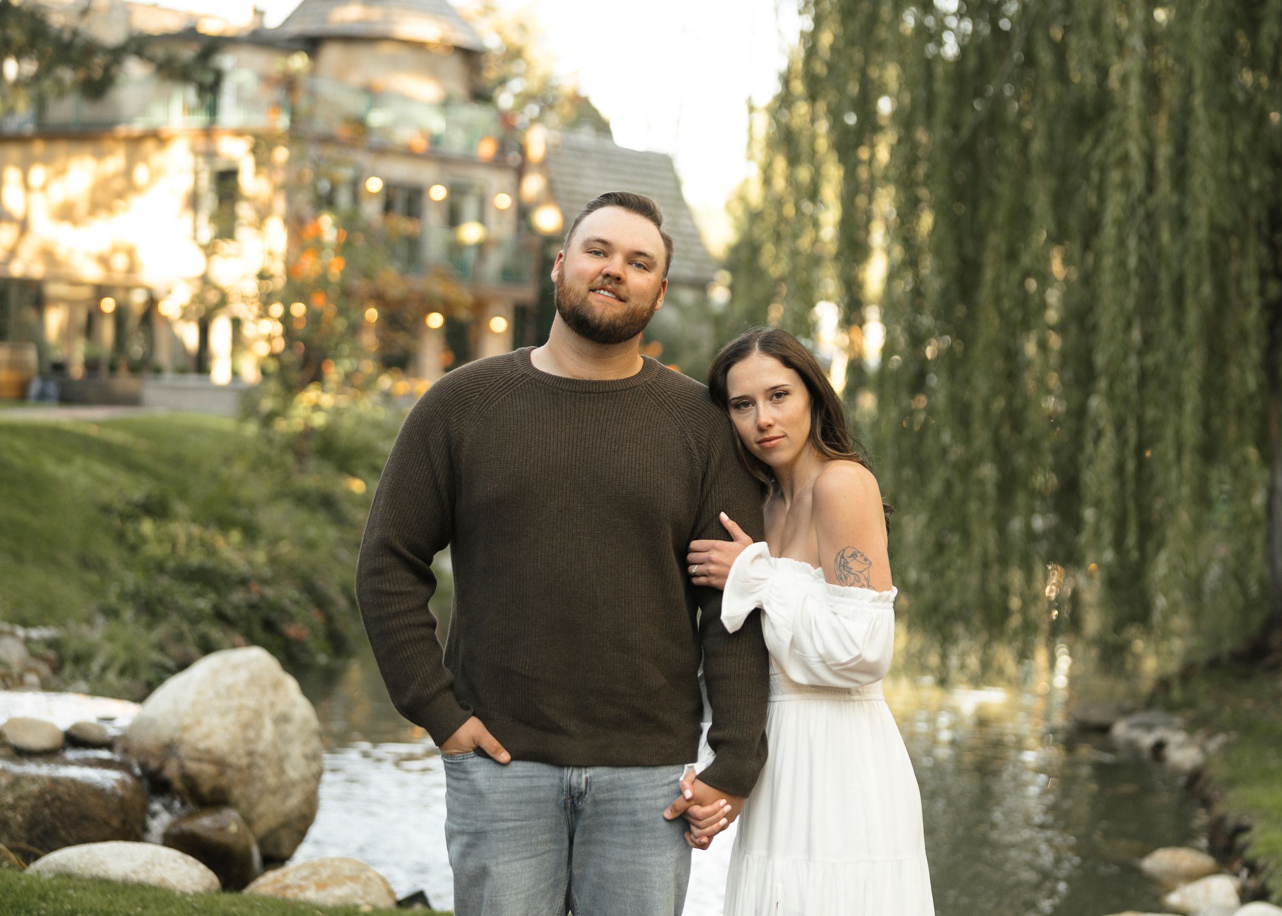 Couple posing outdoors near a creek with willow trees and a building with string lights in the background.