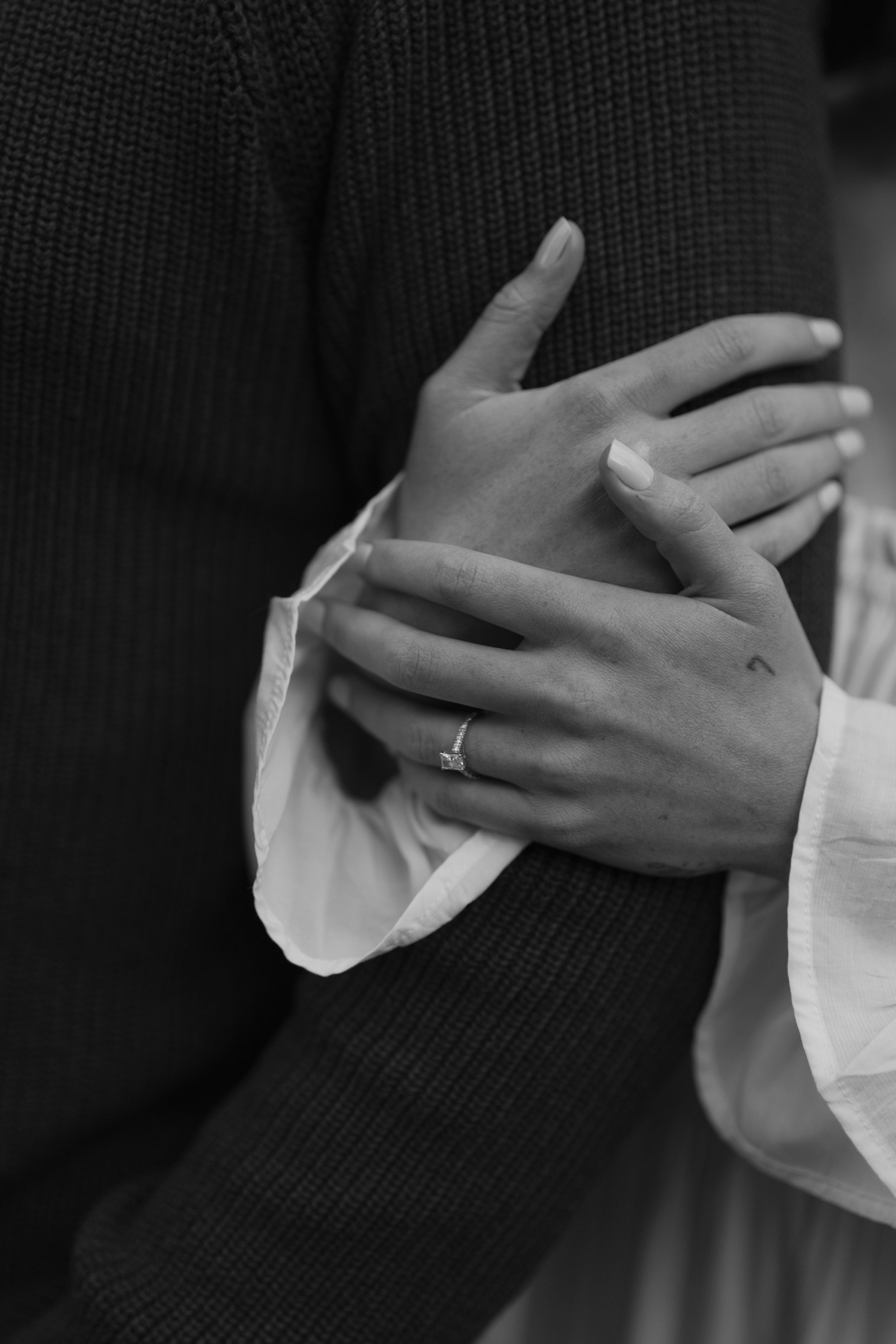 Black and white photo of couple's hands clasped together, woman wearing a diamond engagement ring.