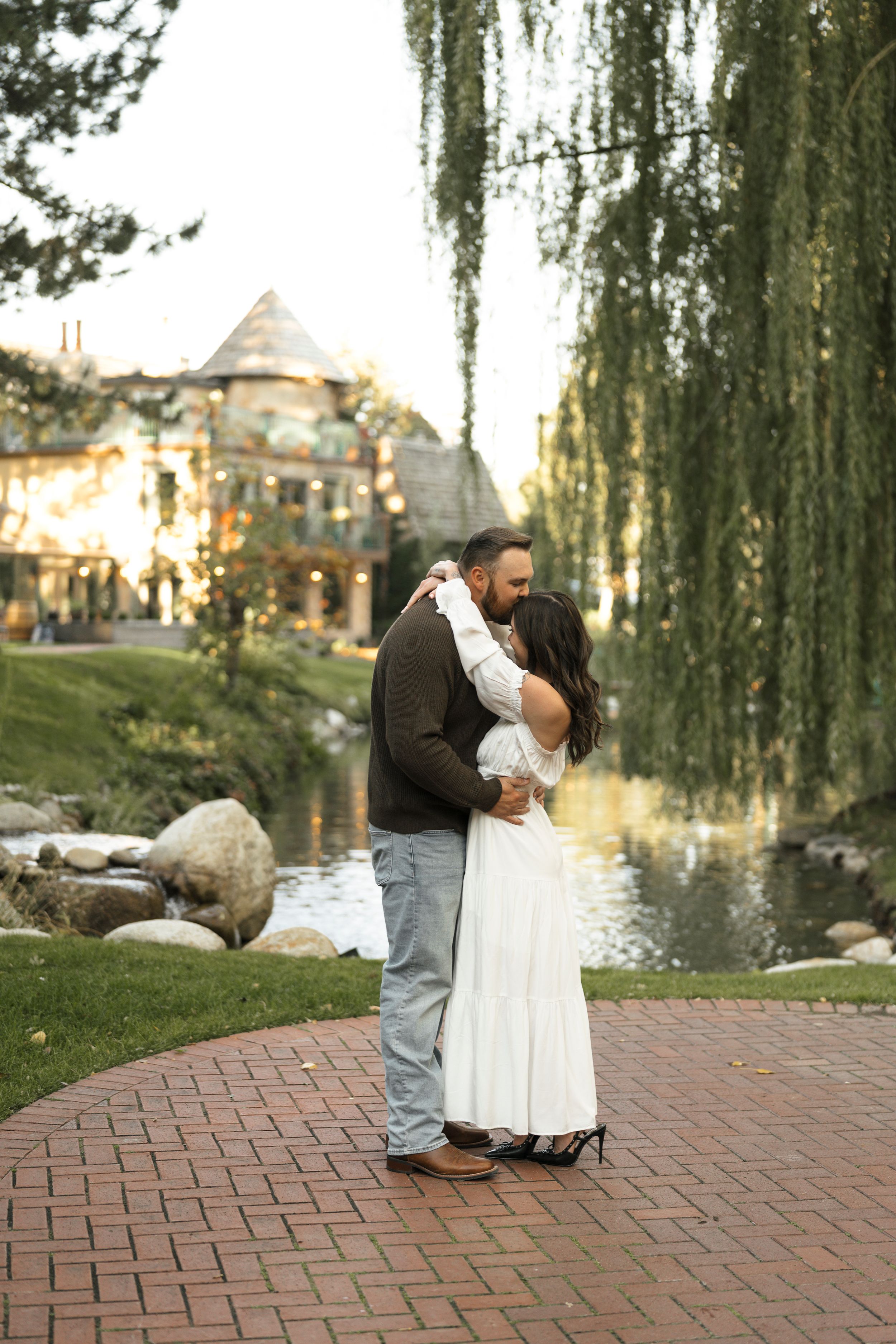 Couple embracing and kissing near a pond with a willow tree and lit venue in background on brick path.