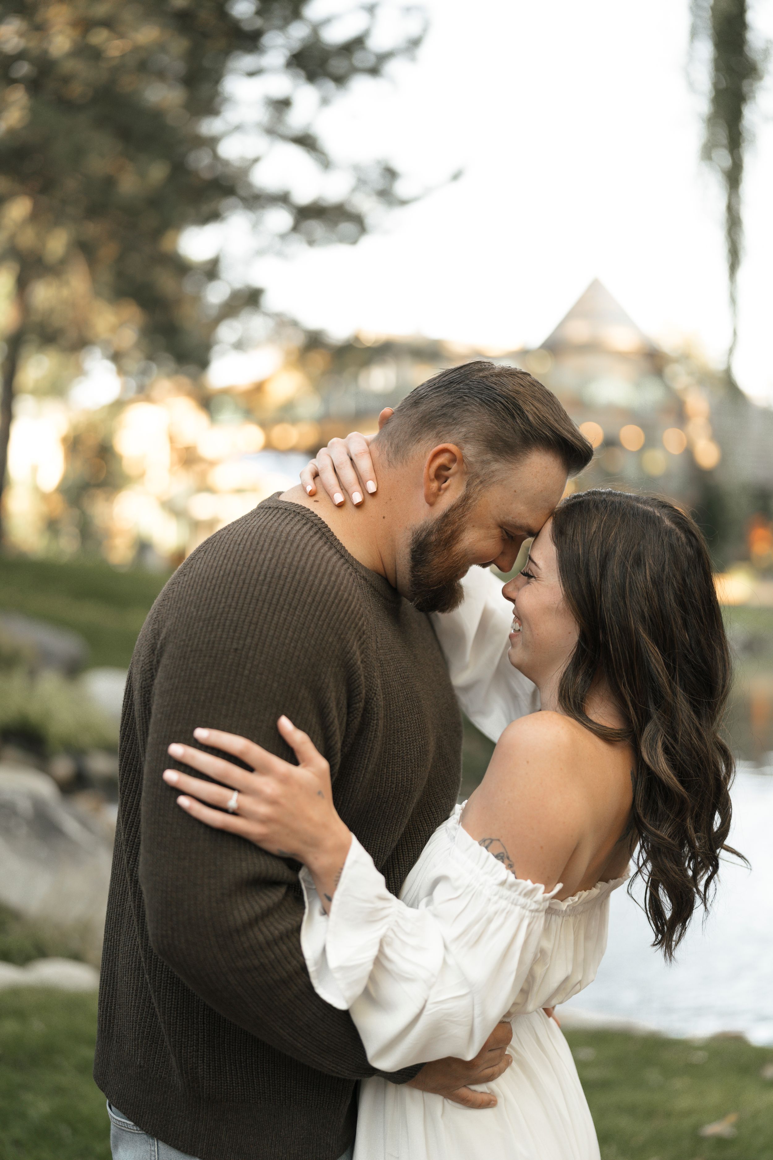 Couple embracing outdoors at golden hour, woman in white off-shoulder dress, man in brown sweater, foreheads touching romantically.