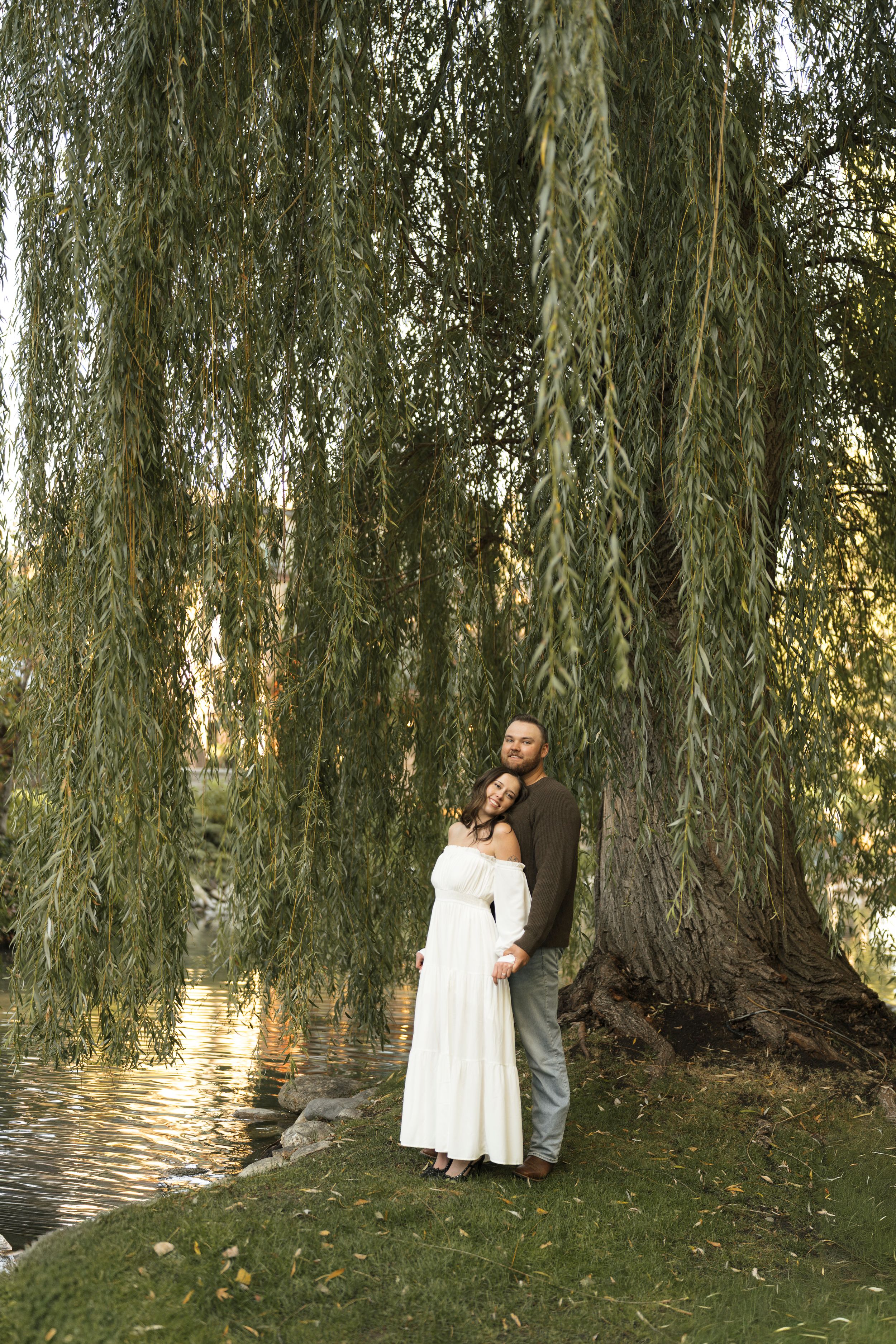 Couple embracing under a weeping willow tree by a pond, woman in white dress, maternity photo shoot outdoors.