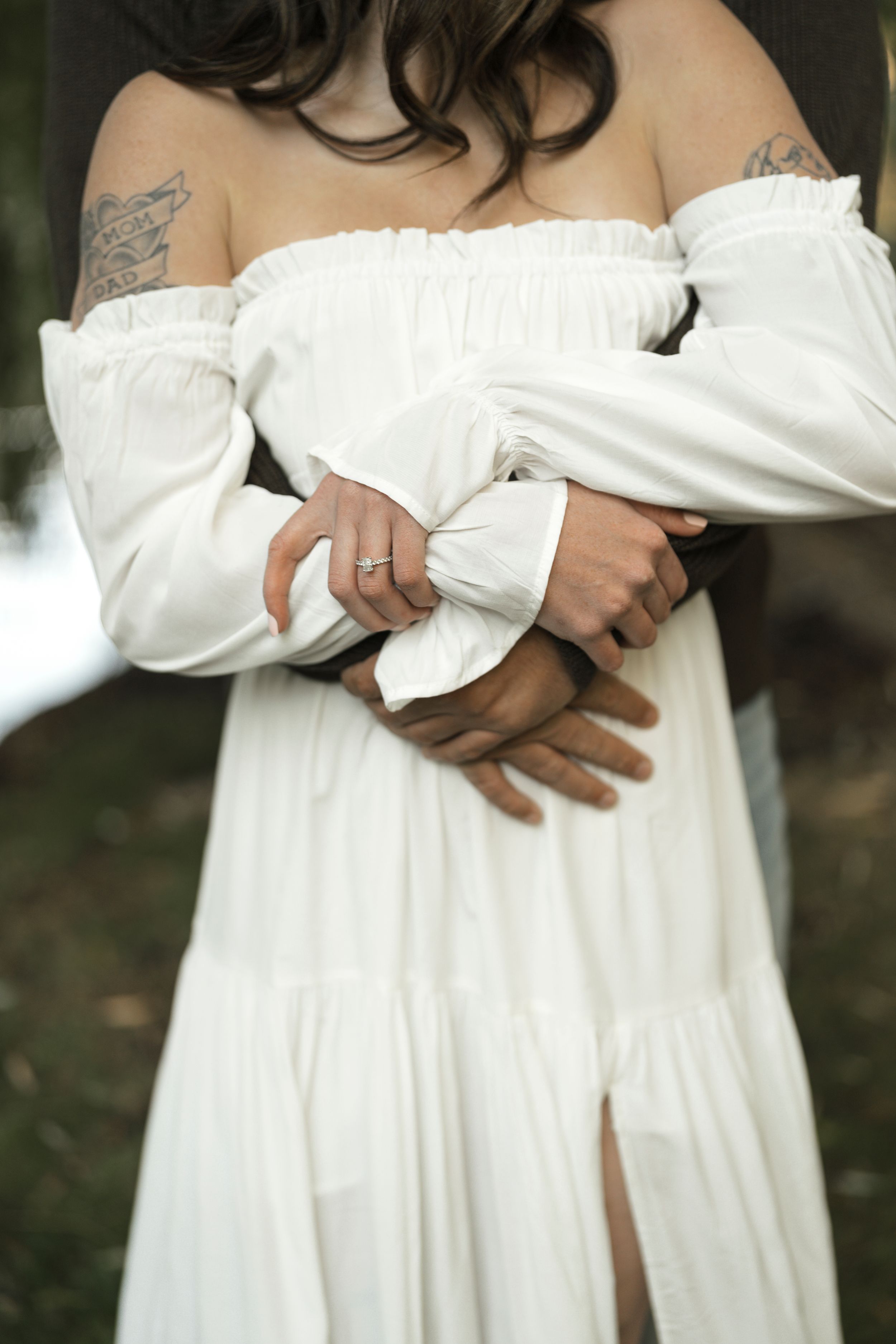 Couple embracing, woman in white off-shoulder dress with tattoos, man's hands wrapped around her waist.