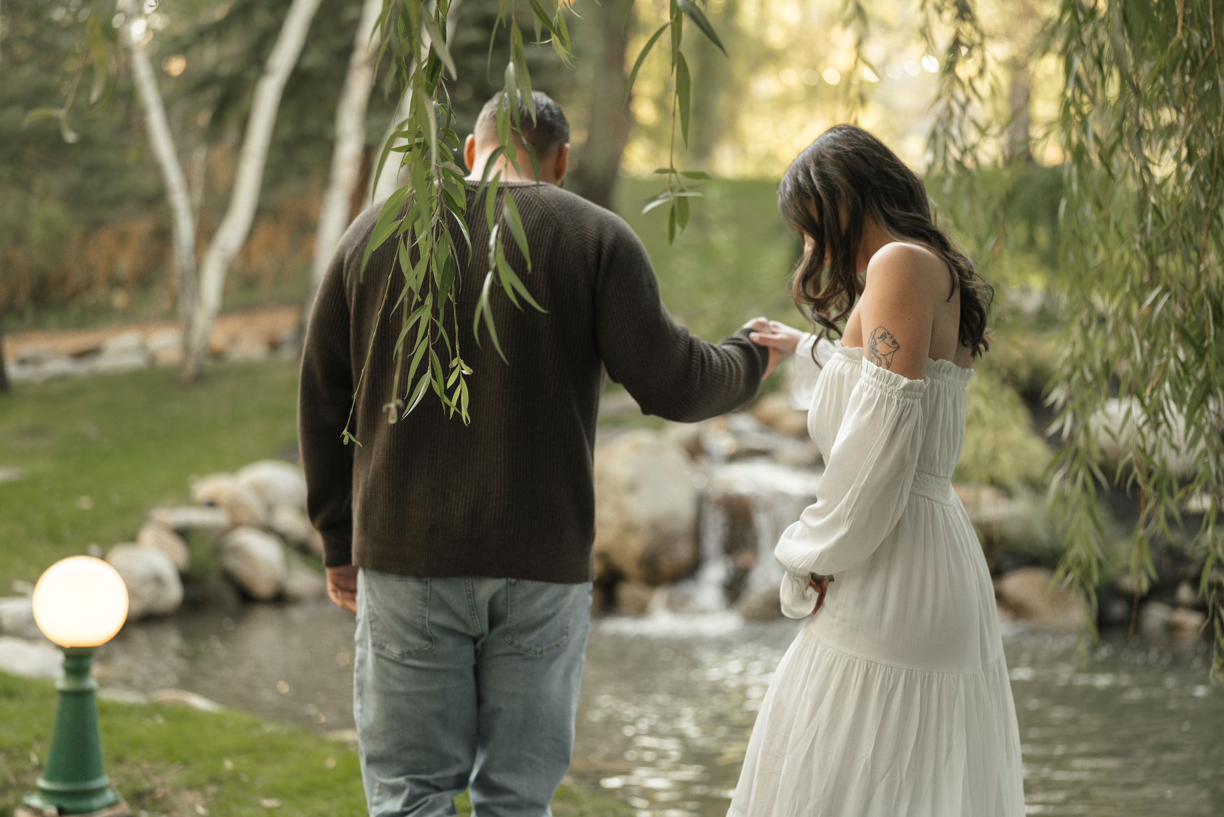 Couple walking hand in hand by a peaceful pond surrounded by weeping willow trees in a lush garden.