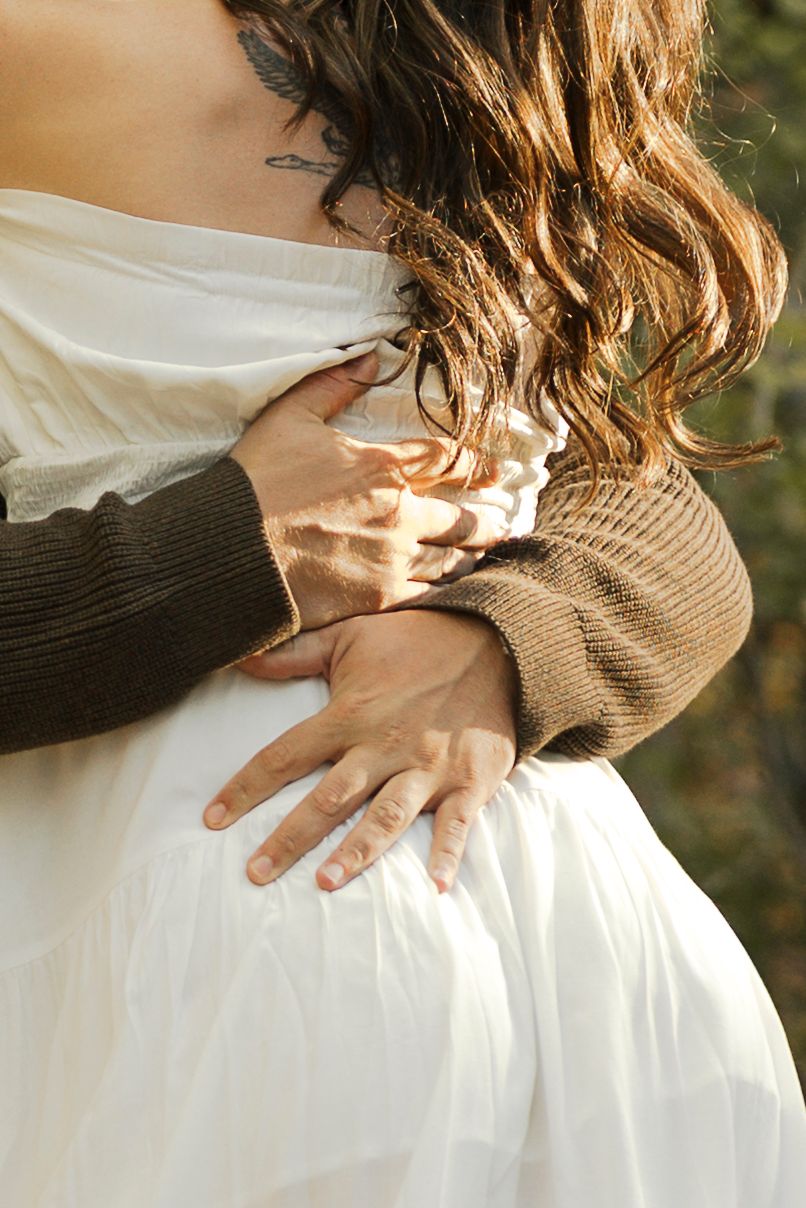 A person in a brown sweater tenderly embraces a pregnant woman in a white dress, cradling her baby bump.