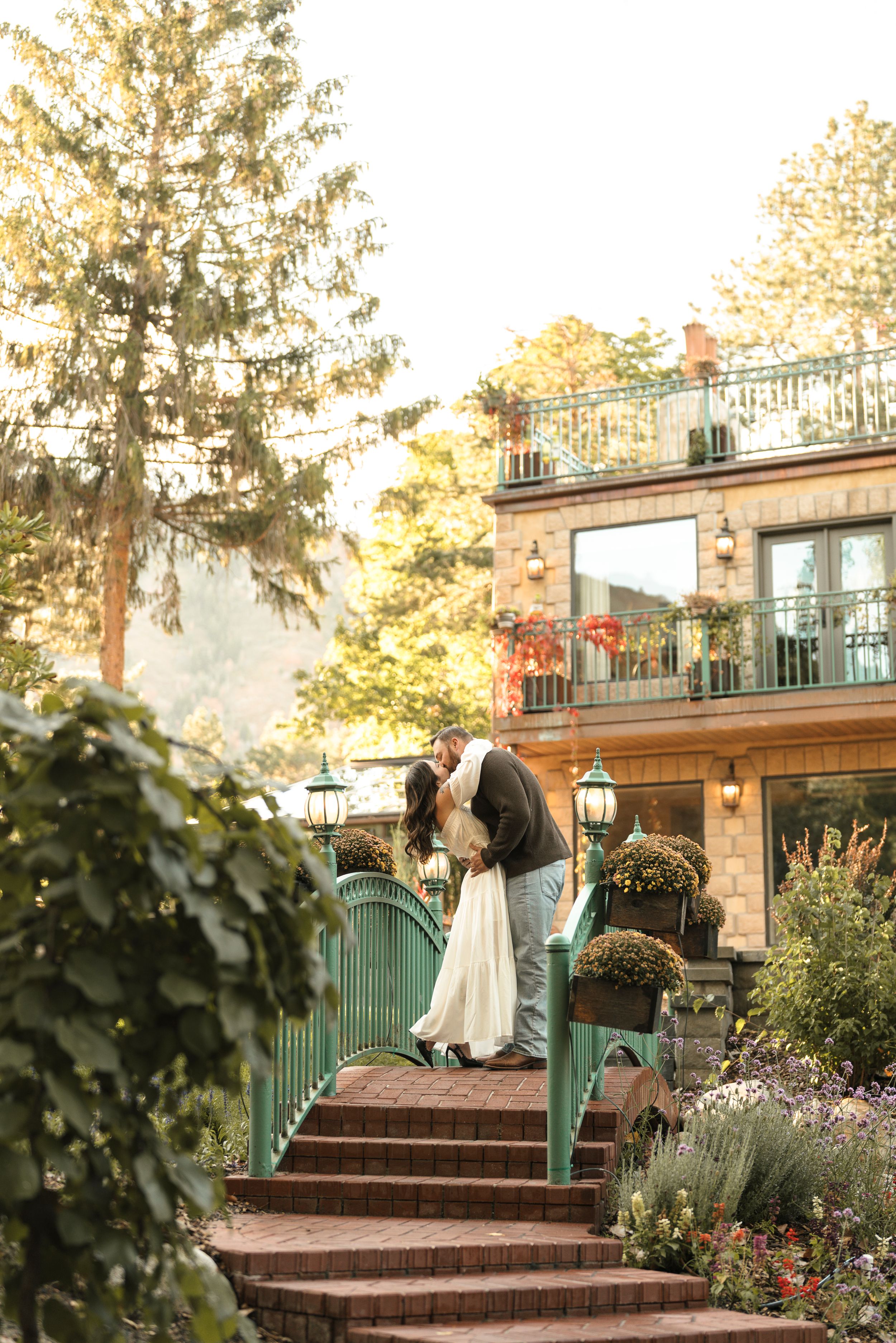 Couple sharing a romantic kiss on brick steps surrounded by lush gardens outside an elegant venue.