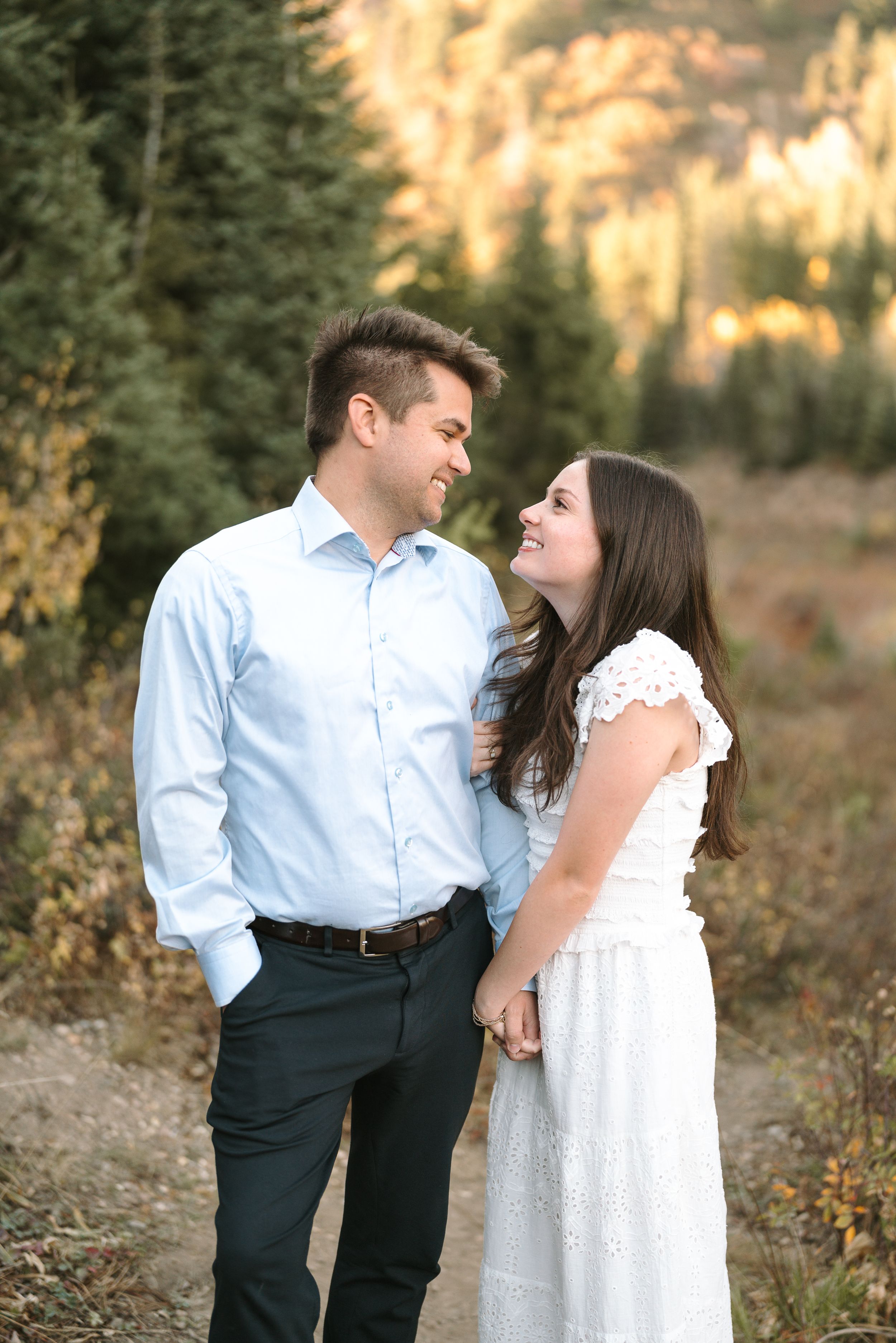 Couple in engagement photos standing together on mountain trail during golden hour.