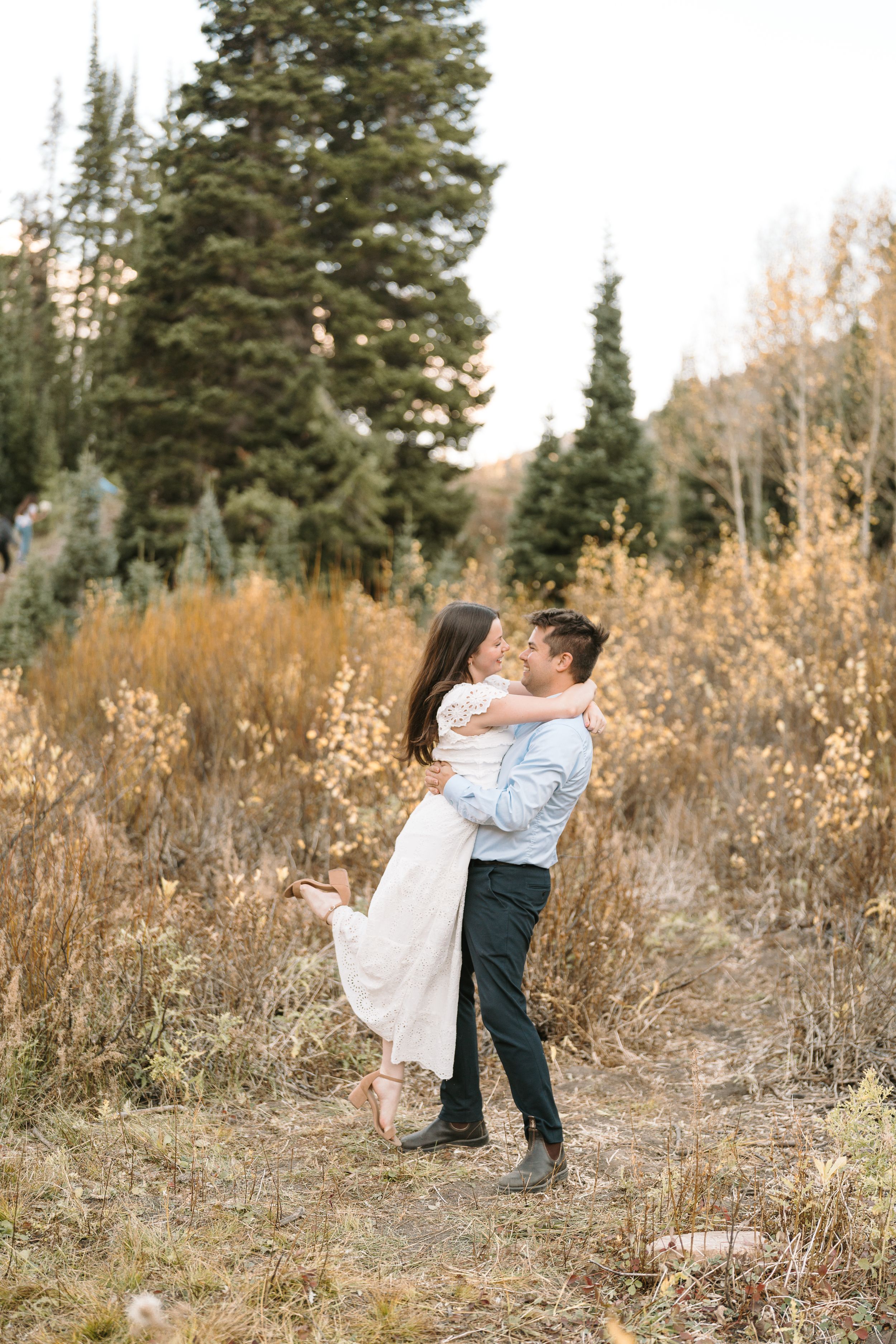 Couple embracing in playful lift pose surrounded by tall grass and evergreen trees.