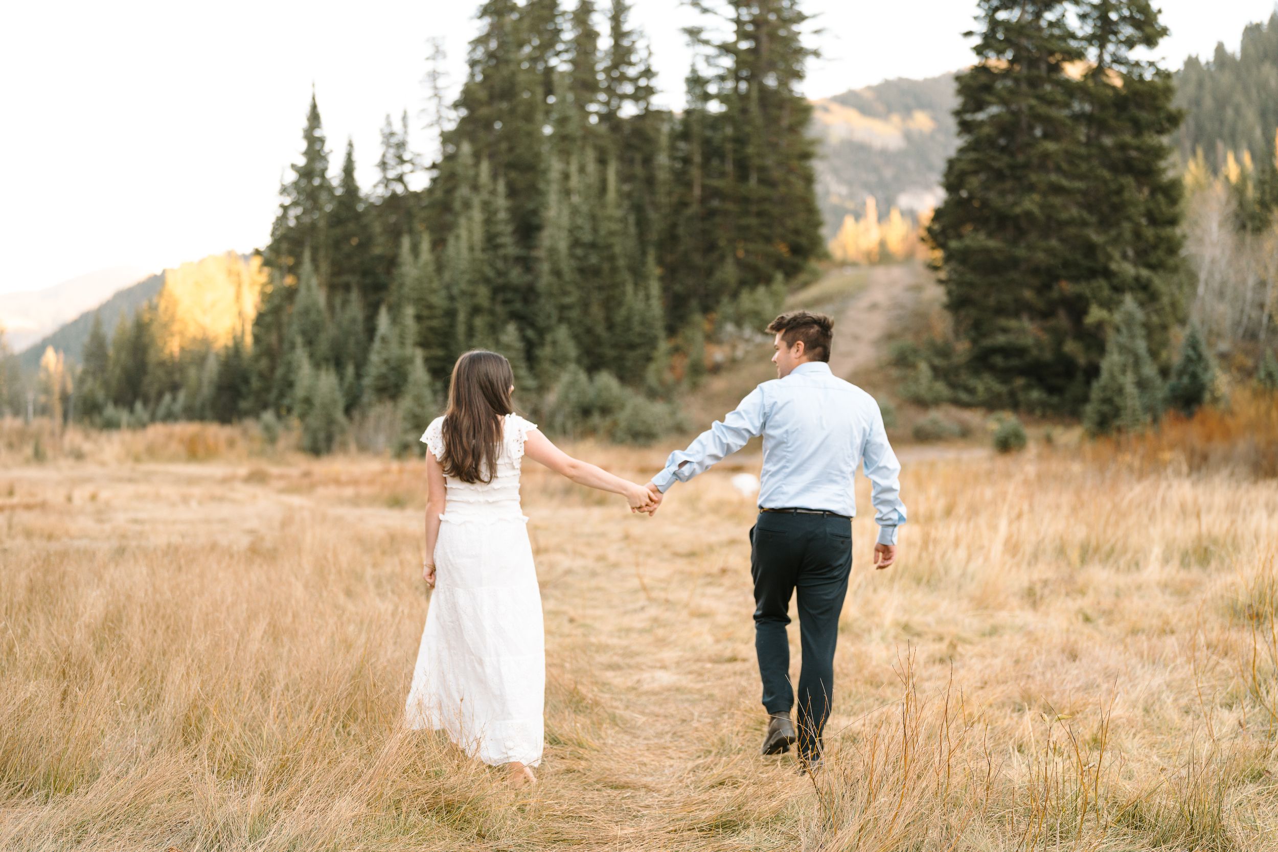 Couple holding hands while walking through mountain meadow during autumn engagement session.