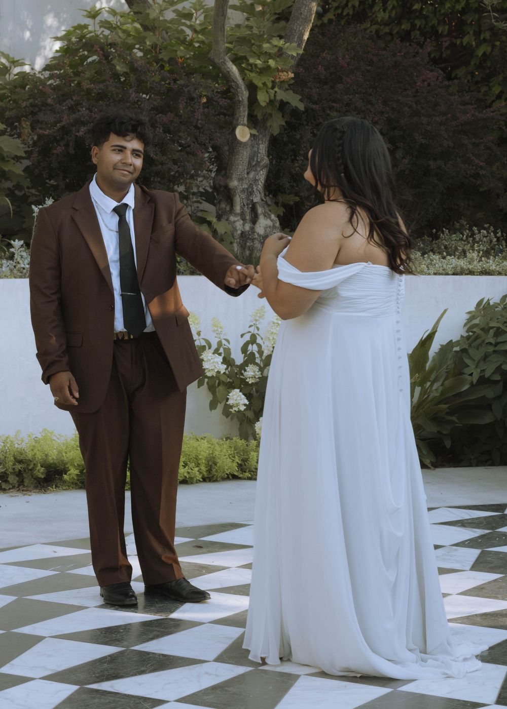 Newlyweds during first dance, groom smiling at bride while taking in her beauty. White flowers and checkered floor in background.