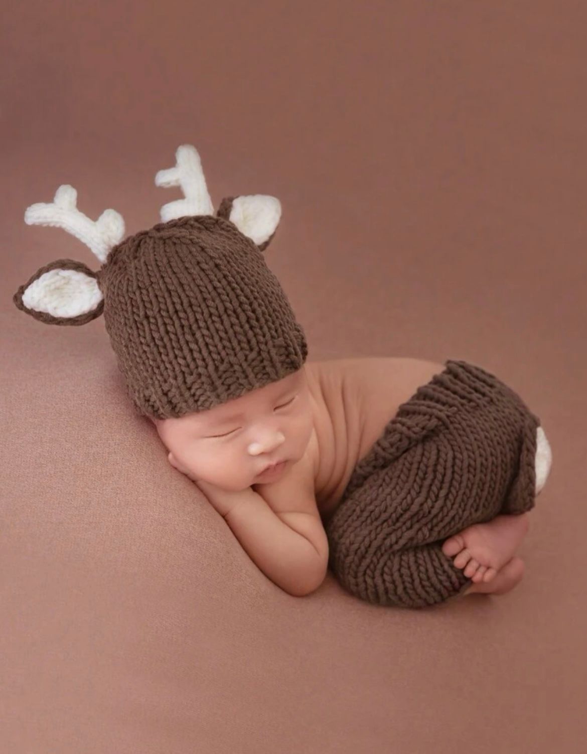 Newborn baby sleeping peacefully wearing a brown knit hat and wrap on brown background.