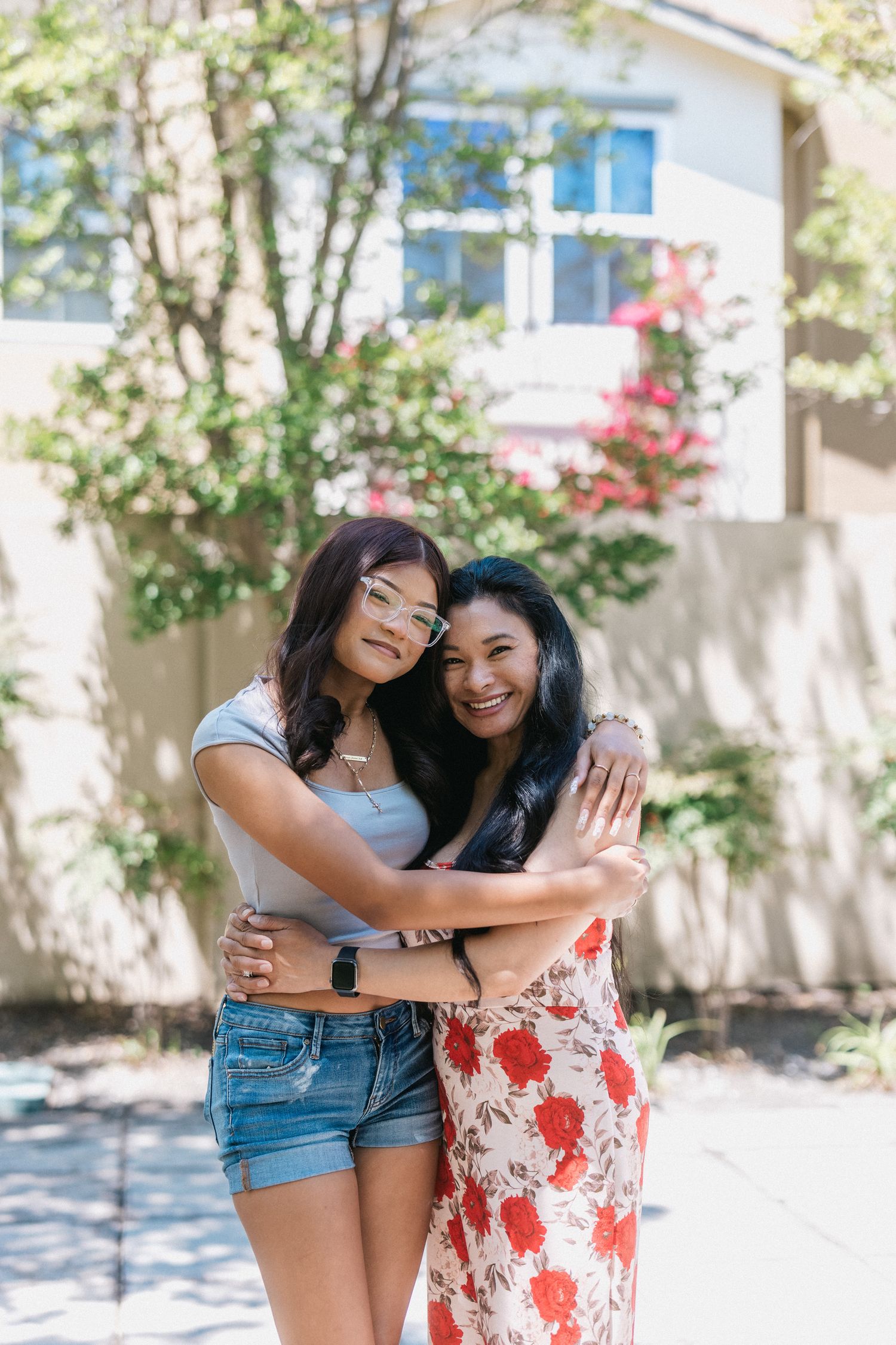 Two friends embrace and smile while standing in front of a white building on a sunny day.
