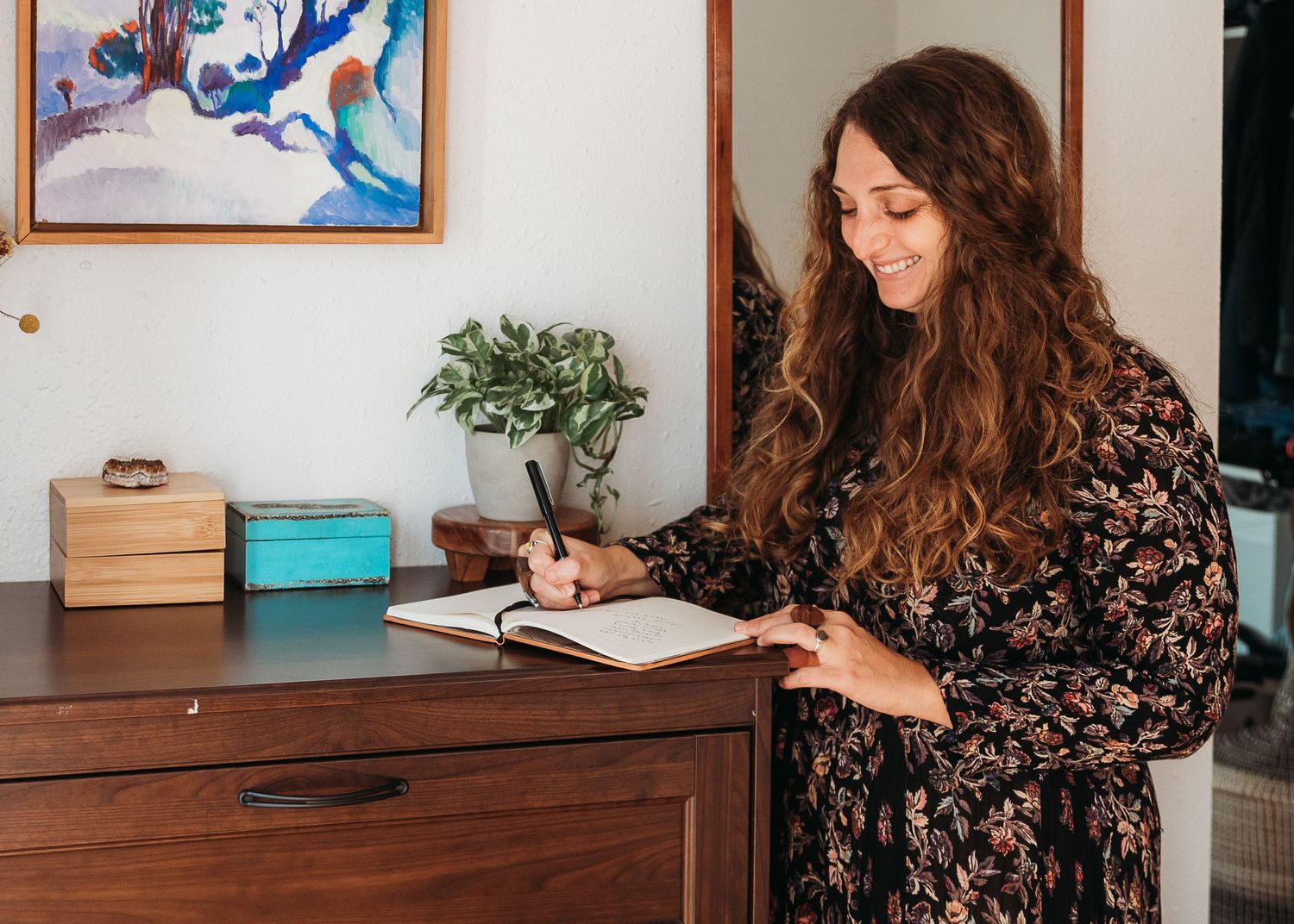 A woman in a floral dress writes in a journal at a wooden desk with plants and decor in the background.