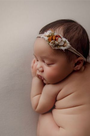 Sleeping newborn baby girl wearing a floral headband, resting peacefully with hands tucked under her cheek.