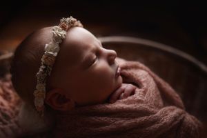 Sleeping newborn baby girl wrapped in dusty rose knit swaddle wearing floral headband in wooden bowl.