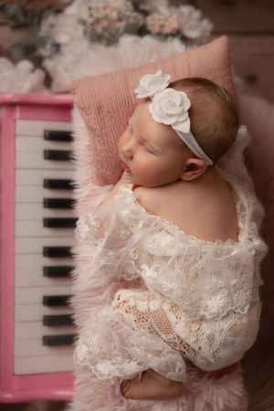 Sleeping newborn baby in white lace outfit and floral headband resting on pink fur beside a pink toy piano.