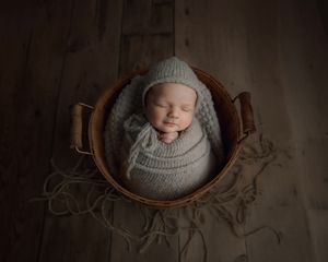 Newborn baby sleeping peacefully in a wicker basket, wrapped in a soft gray knit swaddle and matching bonnet.
