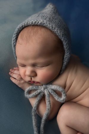 Sleeping newborn baby wearing a gray knit bonnet hat with bow tie, resting peacefully on a blue background.