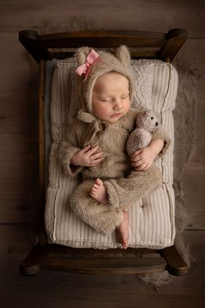Newborn baby sleeping in tiny wooden bed wearing fuzzy bear outfit with pink bow holding matching teddy bear.