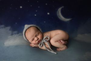 Newborn baby sleeping peacefully wearing a gray knit bonnet on a dreamy blue night sky background with stars and moon.