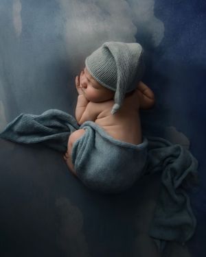 Newborn baby sleeping peacefully wrapped in a blue knit blanket and hat on a dreamy blue background.