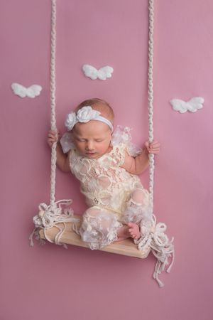 Newborn baby girl in lace outfit sleeping on a wooden swing with white angel wings on pink background.