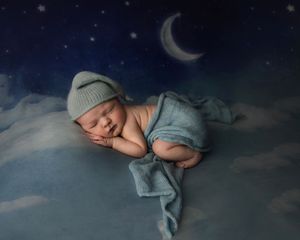 Sleeping newborn baby wrapped in blue, wearing a knit hat, posed on clouds under a crescent moon.