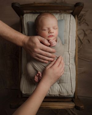 Sleeping newborn swaddled in gray, lying in a wooden cradle with parents hands nearby.