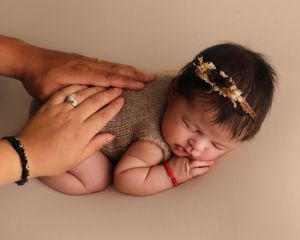 Sleeping newborn baby wearing a floral headband and knit outfit, cradled gently by parent hands on a neutral background.